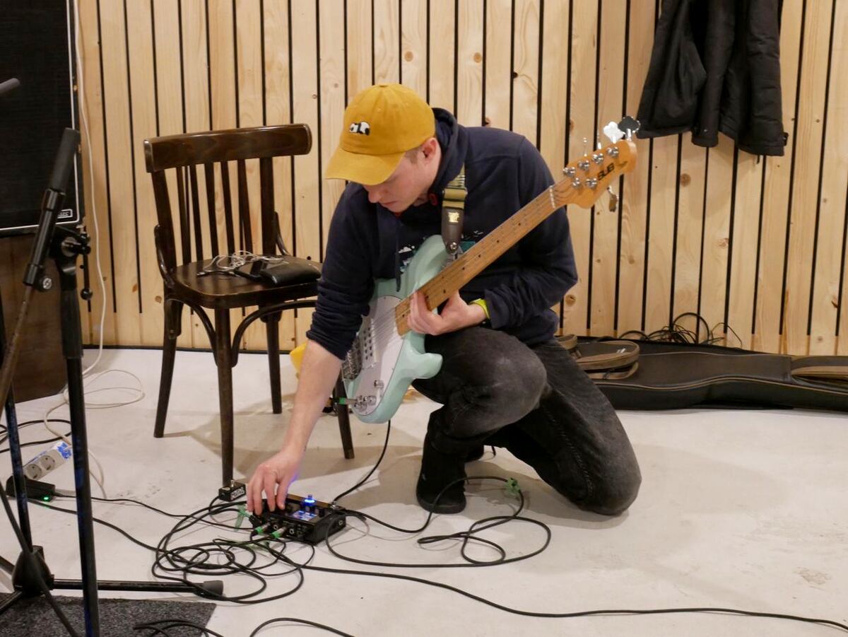 Nandi kneeling on a rehearsal room floor with a mint green bass, adjusting a pedal
